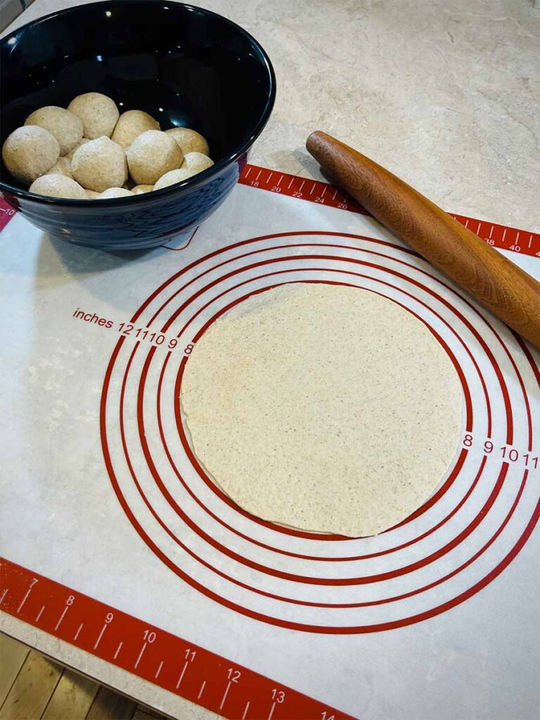 fresh milled flour tortilla dough rolled into a circle on a pastry mat with dough balls in a bowl