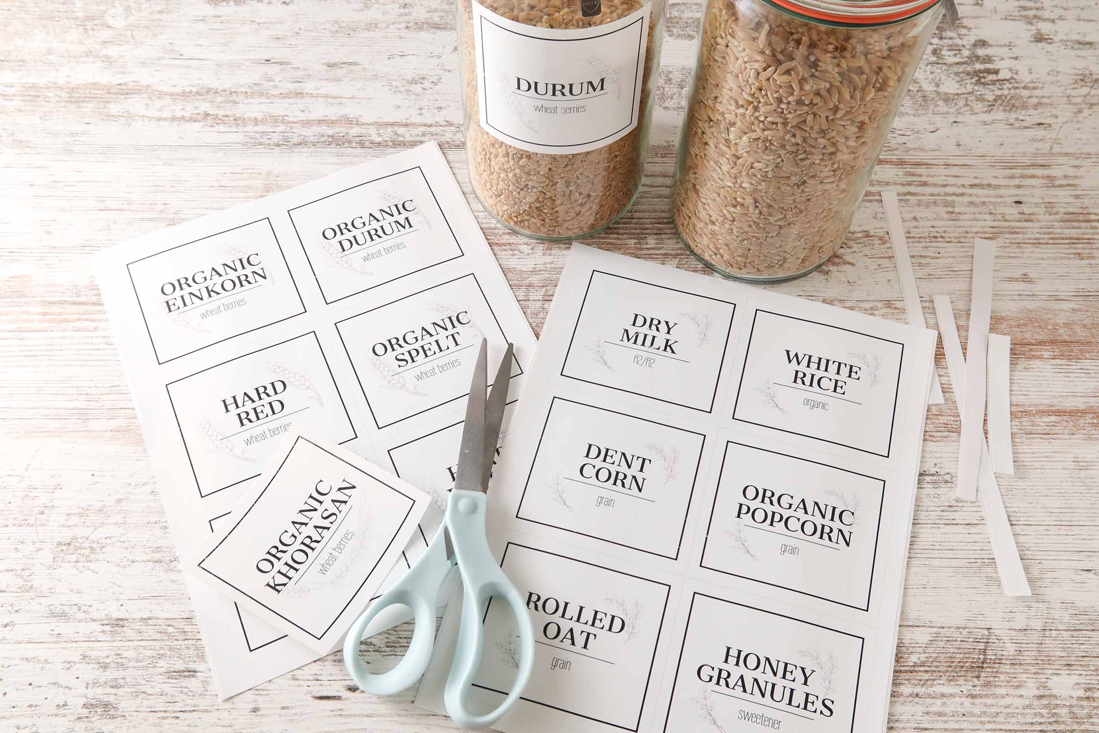 Printable type-your-own-text pantry labels on paper sheets with scissors, cut-out labels, and glass jars of wheat berries.