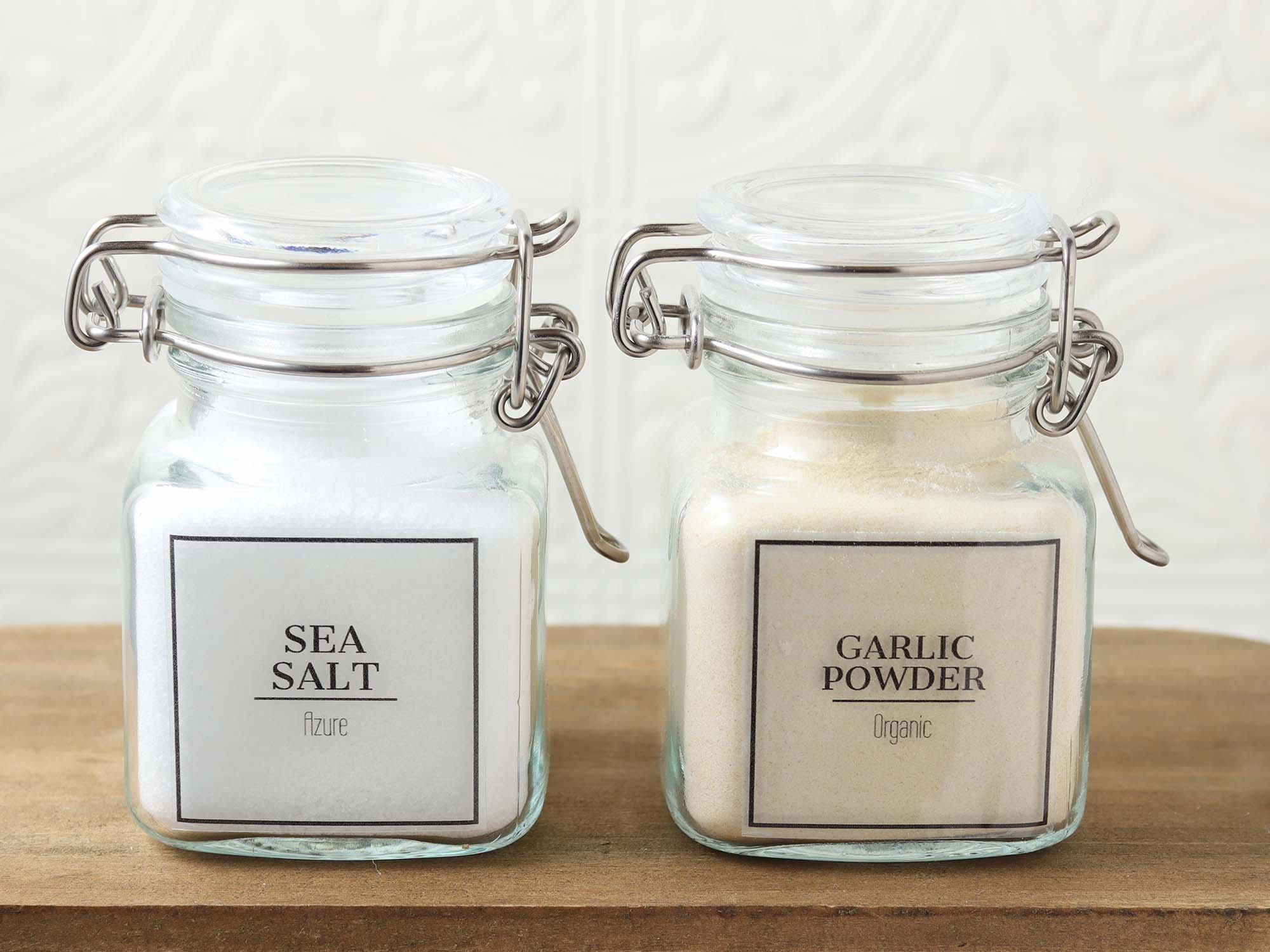Two glass spice jars with type-your-own-text pantry labels printed on clear sticker paper, featuring sea salt and garlic powder.